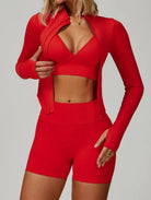 Front view of a woman wearing the ContourFlex Three-Piece Set in red, featuring a sports bra, fitted jacket, and shorts, posing with one hand adjusting the jacket.