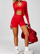 Woman wearing the ContourFlex Three-Piece Set in red, featuring a crop top, fitted jacket, and shorts, posing with one leg forward while holding a matching red gym bag.